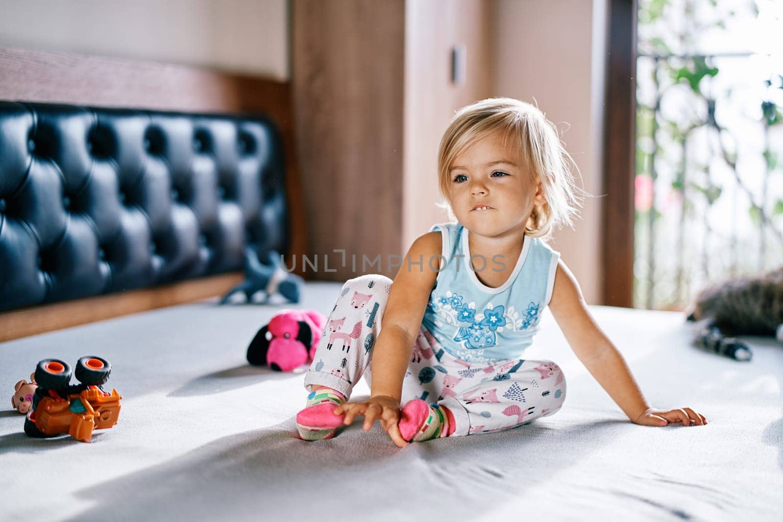 Little girl sitting on the bed surrounded by toys by Nadtochiy