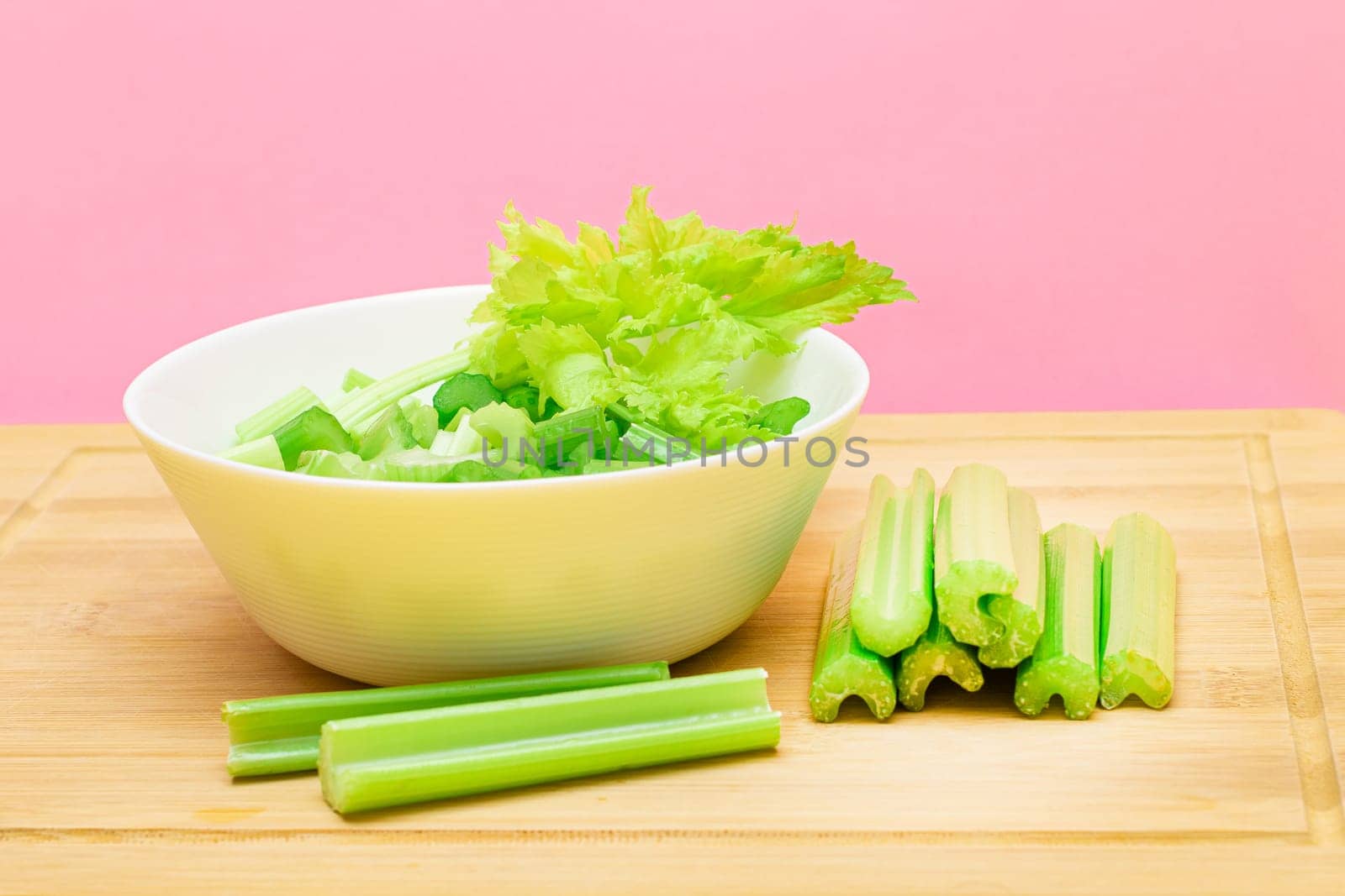 Fresh Chopped Celery Slices in White Bowl with Celery Sticks on Bamboo Cutting Board by InfinitumProdux