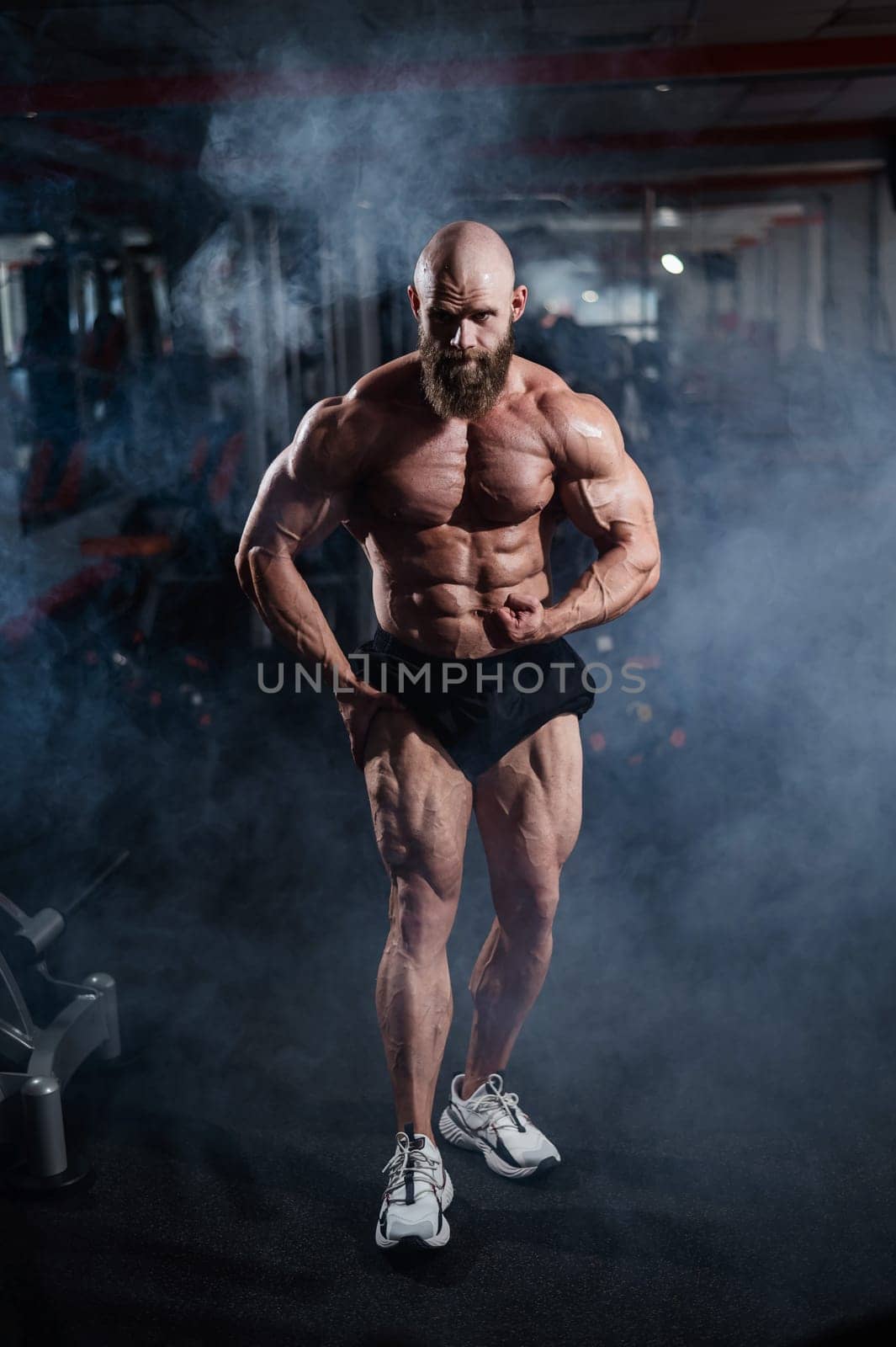 Muscular bald man posing in shorts. Bodybuilder showing off his shape in the gym. by mrwed54