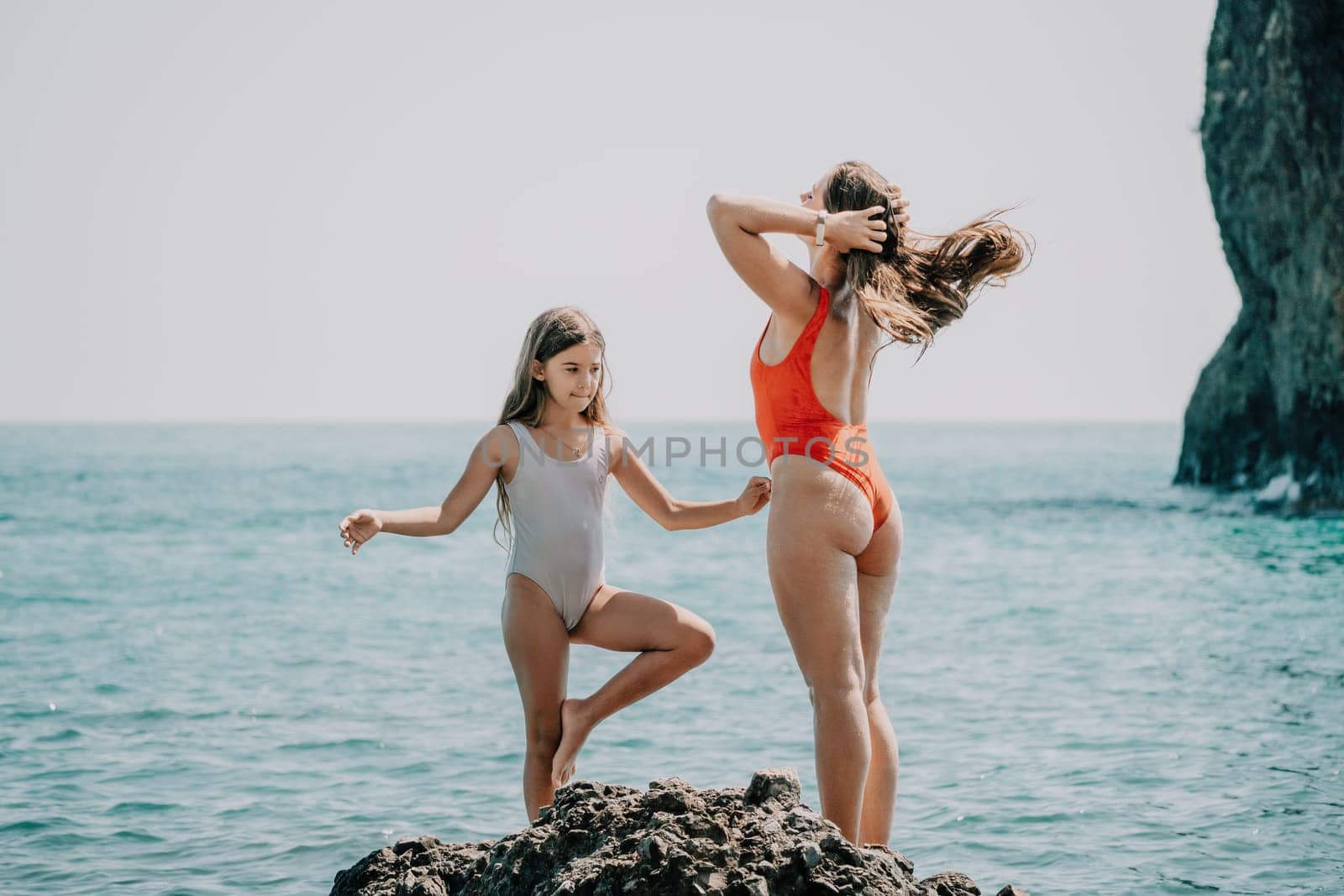 Woman and her daughter practicing balancing yoga pose on one leg up together on rock in the sea. Silhouette mother and daughter doing yoga at beach by panophotograph