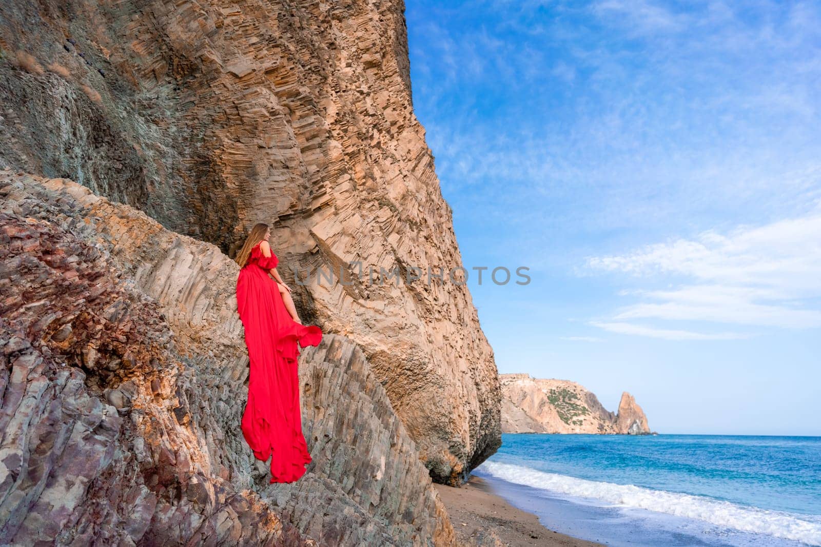 woman sea red dress. Woman with long hair on a sunny seashore in a red flowing dress, back view, silk fabric waving in the wind. Against the backdrop of the blue sky and mountains on the seashore. by Matiunina