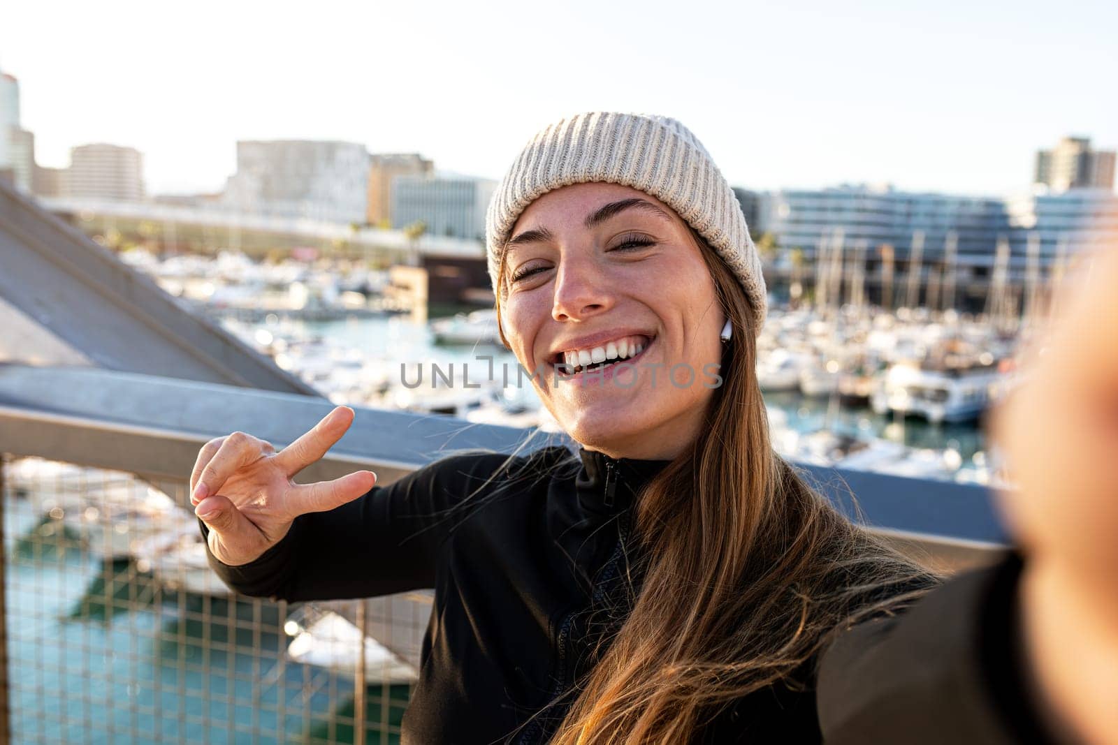 Young happy caucasian jogger woman taking selfie while running outdoors during winter in the city. Female runner wearing sports clothing and hat looking at camera. Sports and technology concept.