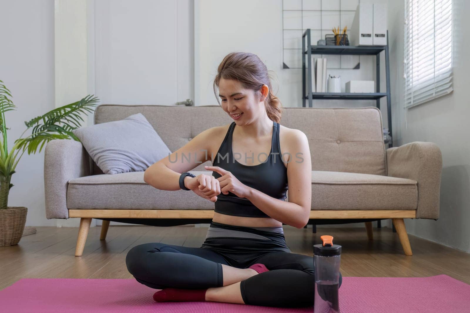 Young fit Asian woman sits on the floor and looks at her smart watch in the living room. An athlete is viewing her performance on a fitness smartwatch app after a workout. by wichayada