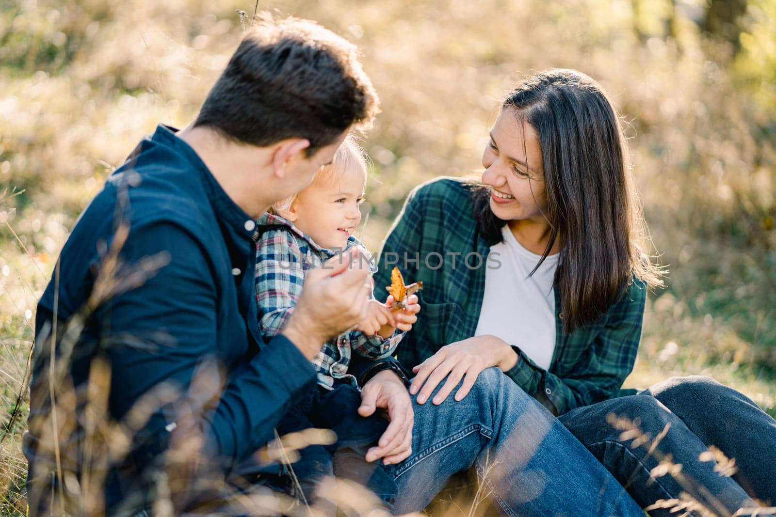 Smiling mom and dad looking at little girl sitting on sunny meadow. High quality photo