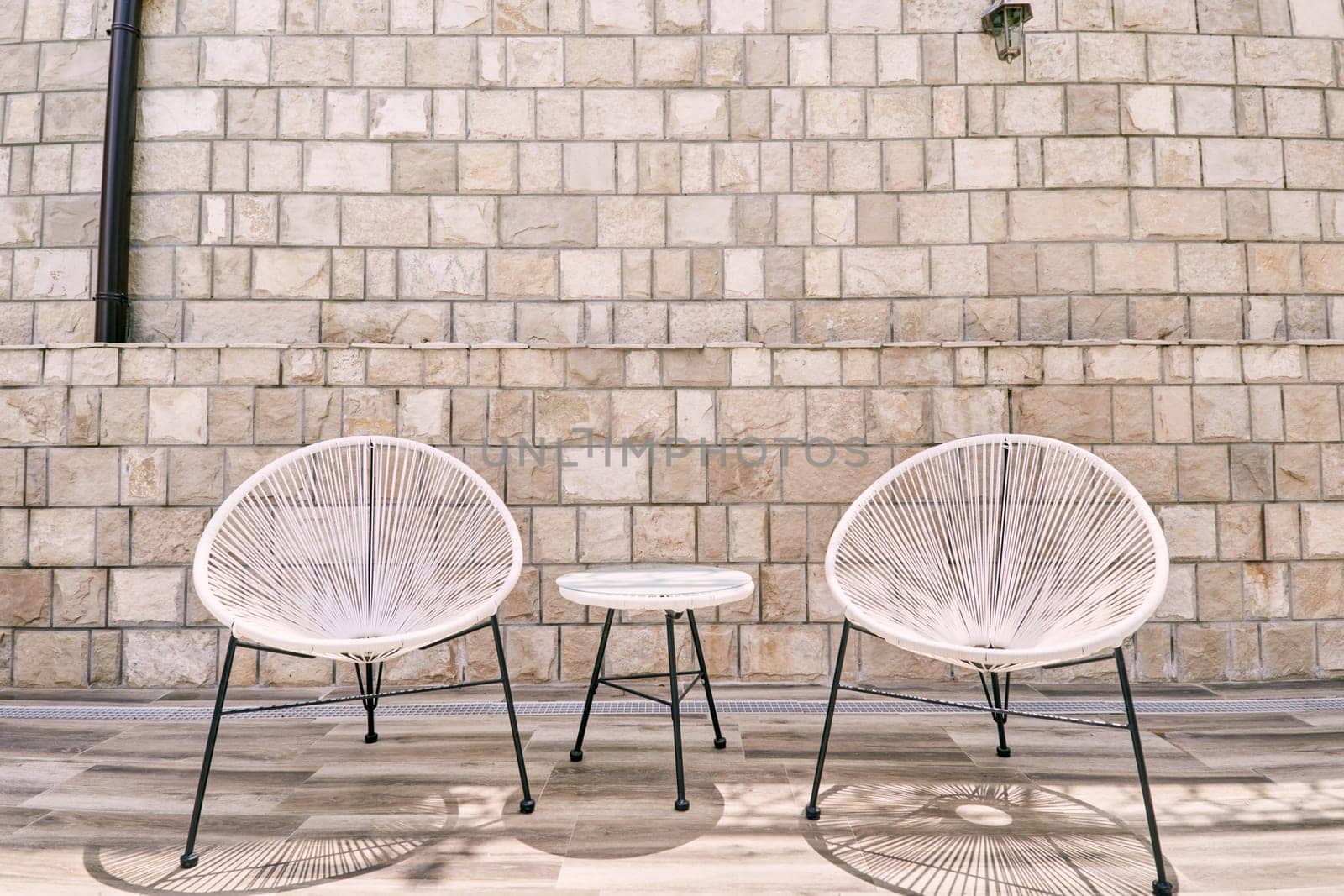 White round polyrattan chairs stand near a table against a stone wall. High quality photo