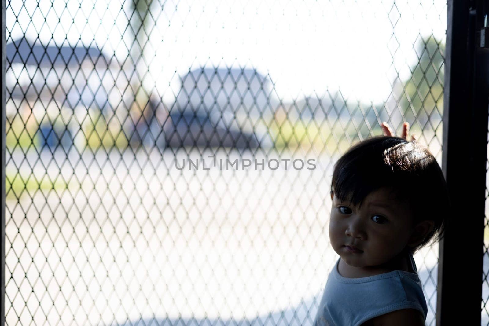 The boy is holding his hands on the fence. The Boy Holds On To The Iron Fence With His Hand. The boy stands behind the fence with emotional expression.
