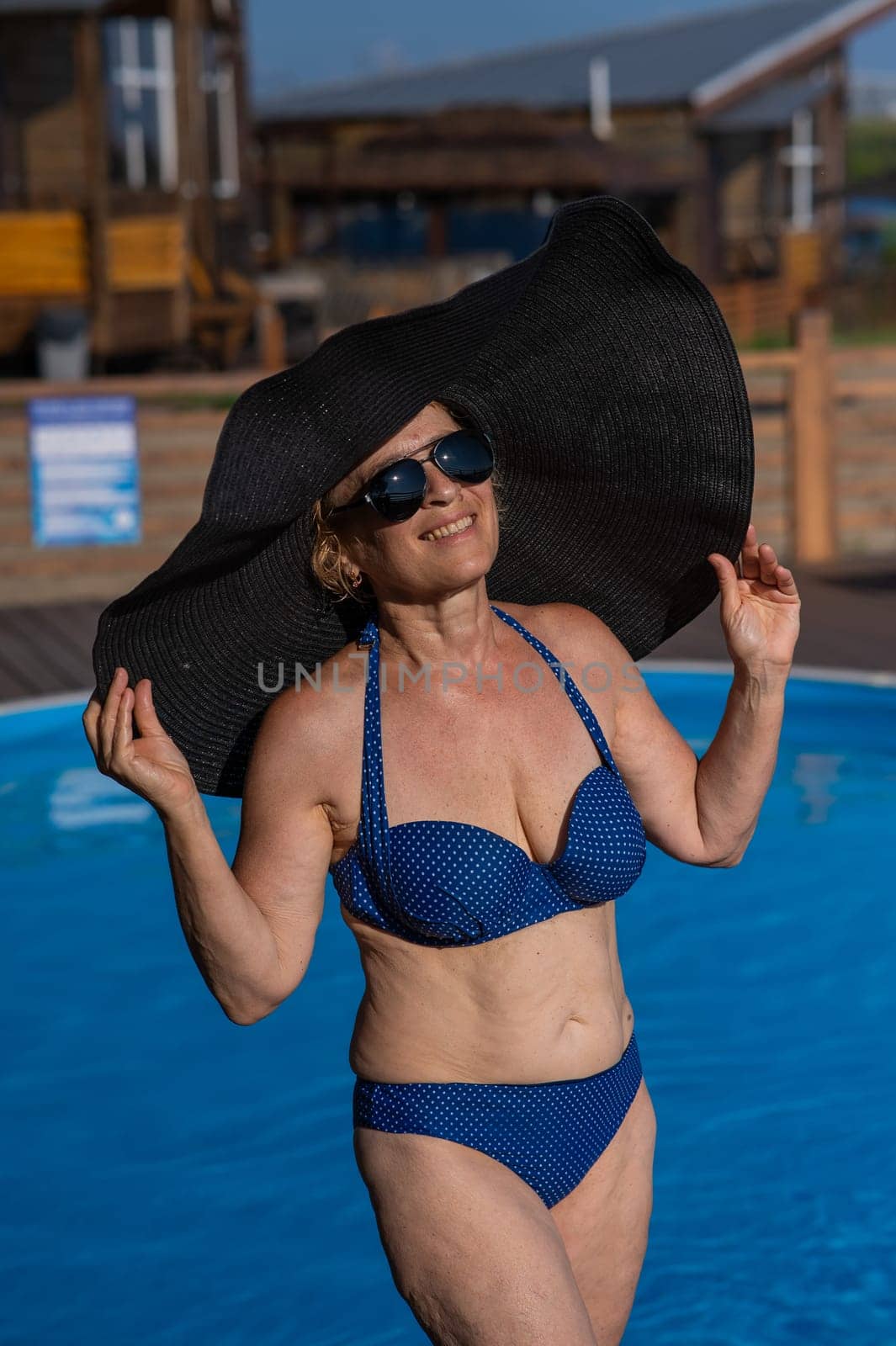 An elderly woman in a huge straw hat and sunglasses is sunbathing by the pool. Retiree on vacation