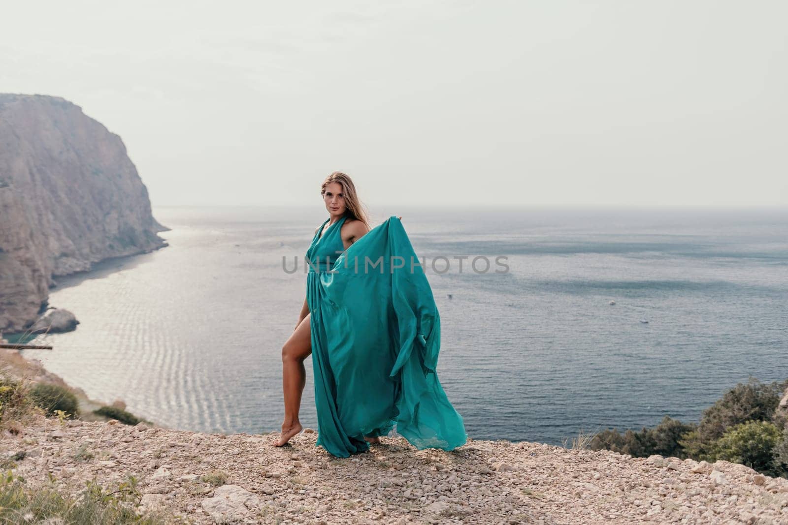 Side view a Young beautiful sensual woman in a mint long dress posing on a volcanic rock high above the sea during sunset. Girl on the nature on overcast sky background. Fashion photo