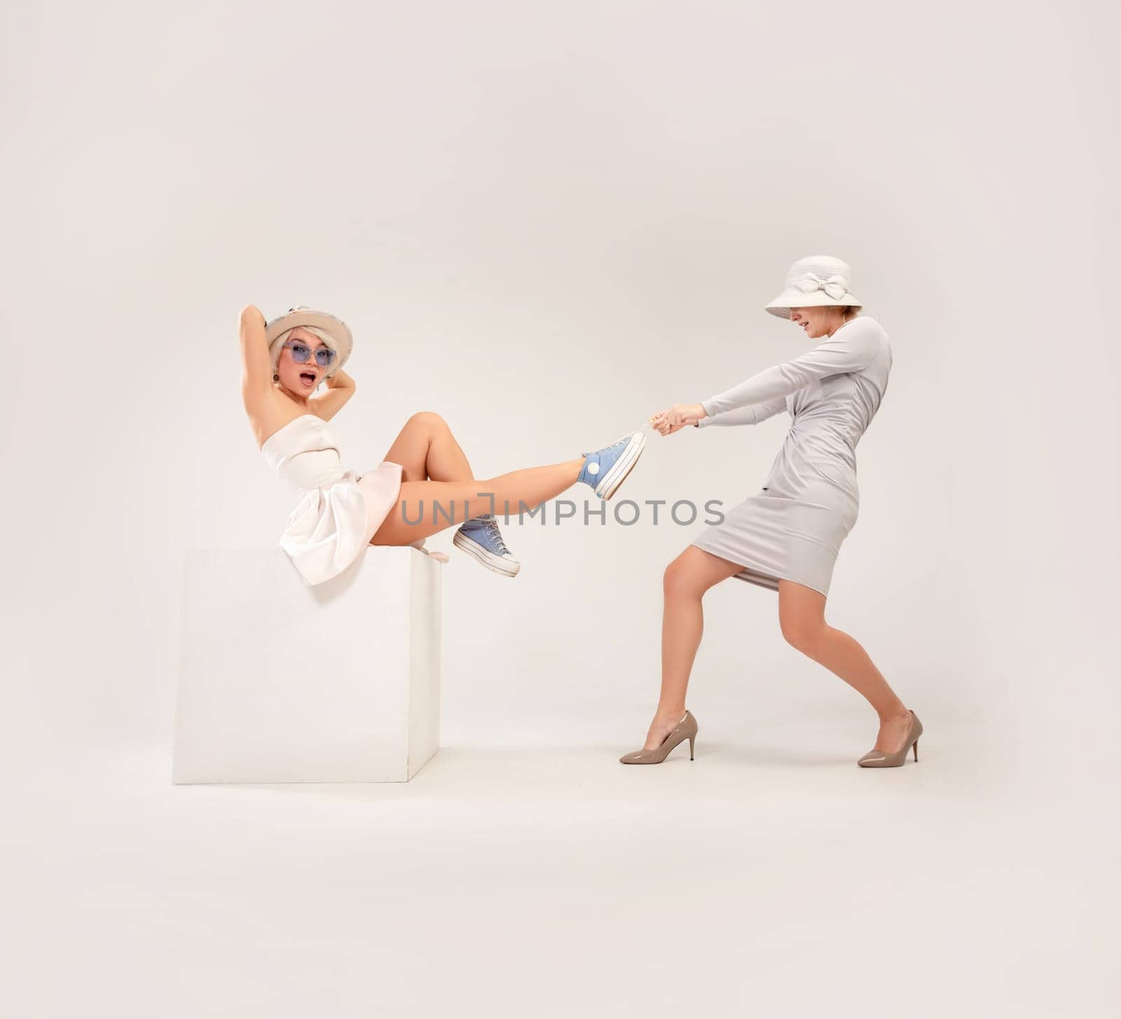 a studio photo of two girls in different styles of clothing, playing out a conflict or quarreling with each other on a white background, one pulling the other's foot while taking away shoes