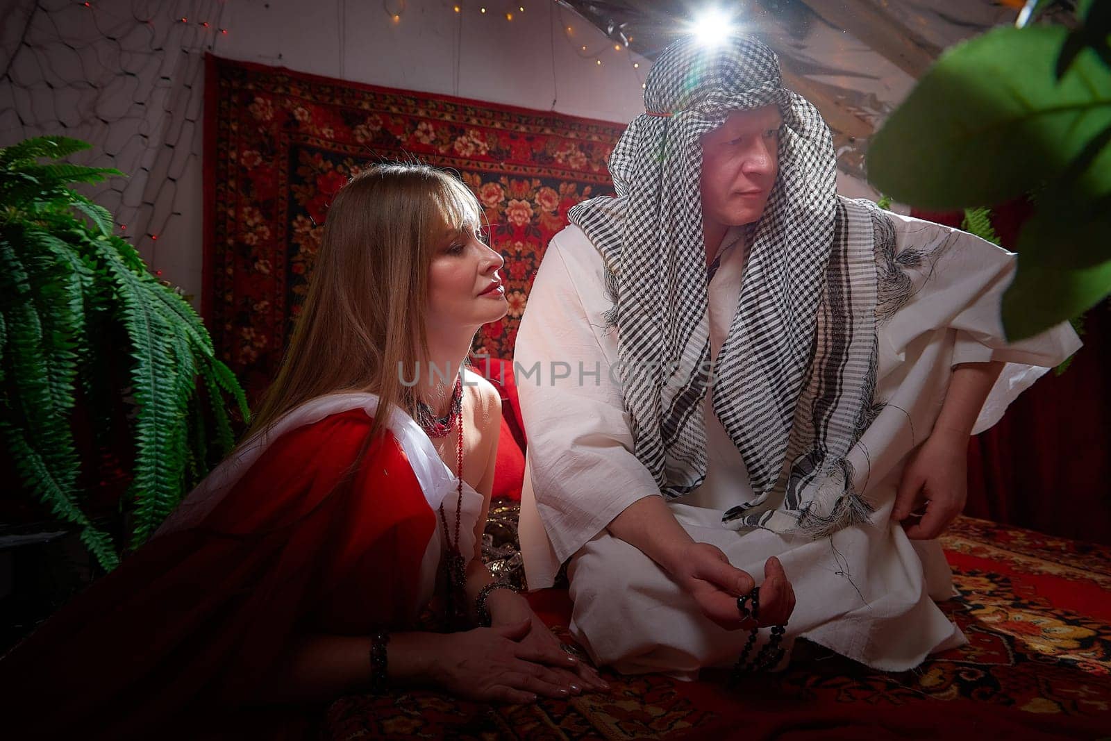 Portrait of young arabian muslim couple in traditional clothes in cozy red room. Fhoto shoot in easten style with male and female model like in harem with a sultan and an odalisque. Partial focus