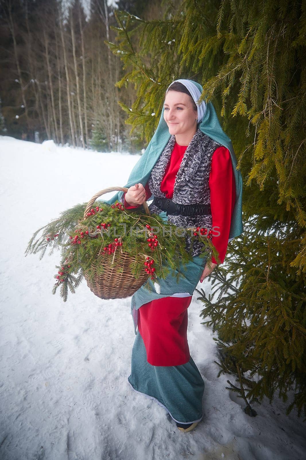 Girl in thick coat and a red sash with basket of fir branches and berries in cold winter day in forest. Medieval peasant woman with firewood. Photoshoot in stile of Christmas fairy tale by keleny