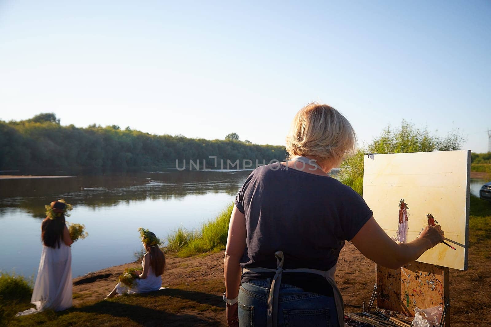 Adult female artist painting picture near water of river or lake in nature and girls in white sundress and flower wreath posing in the holiday of Ivan Kupala in nature at sunset by keleny