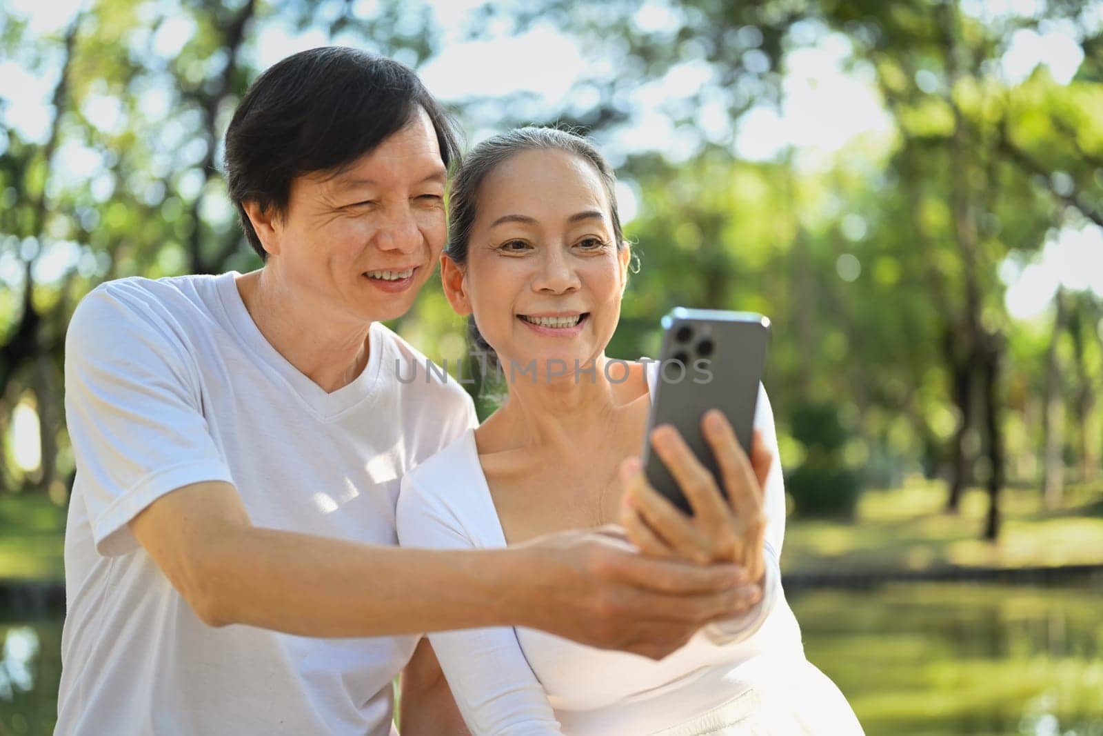 Smiling senior active couple having fun using smart phones while relaxing together in park. by prathanchorruangsak