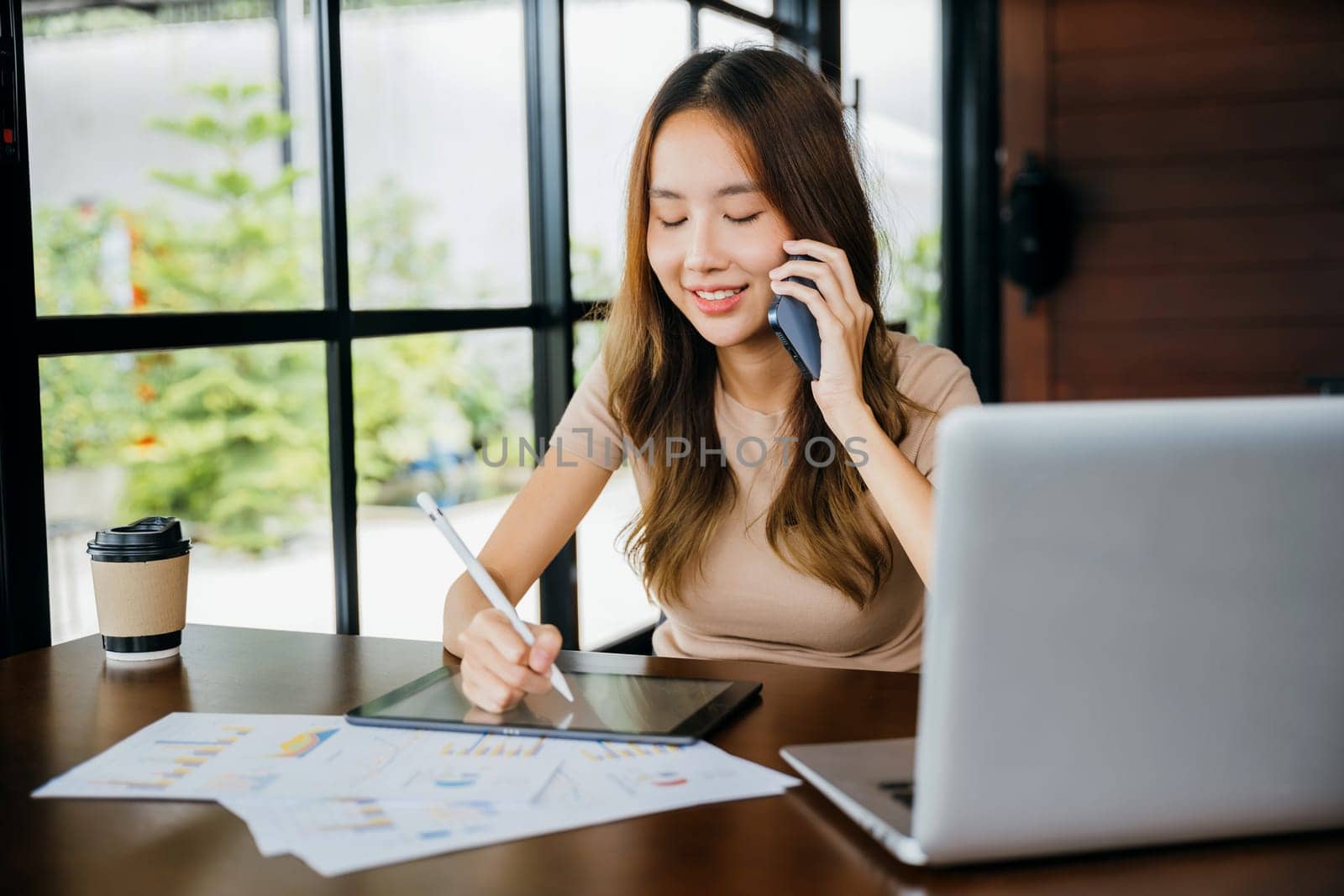 Asian businesswoman sitting at cafe calling mobile phone talking with customer asking about something, smiling female working laptop computer and writing take note on digital tablet at home office