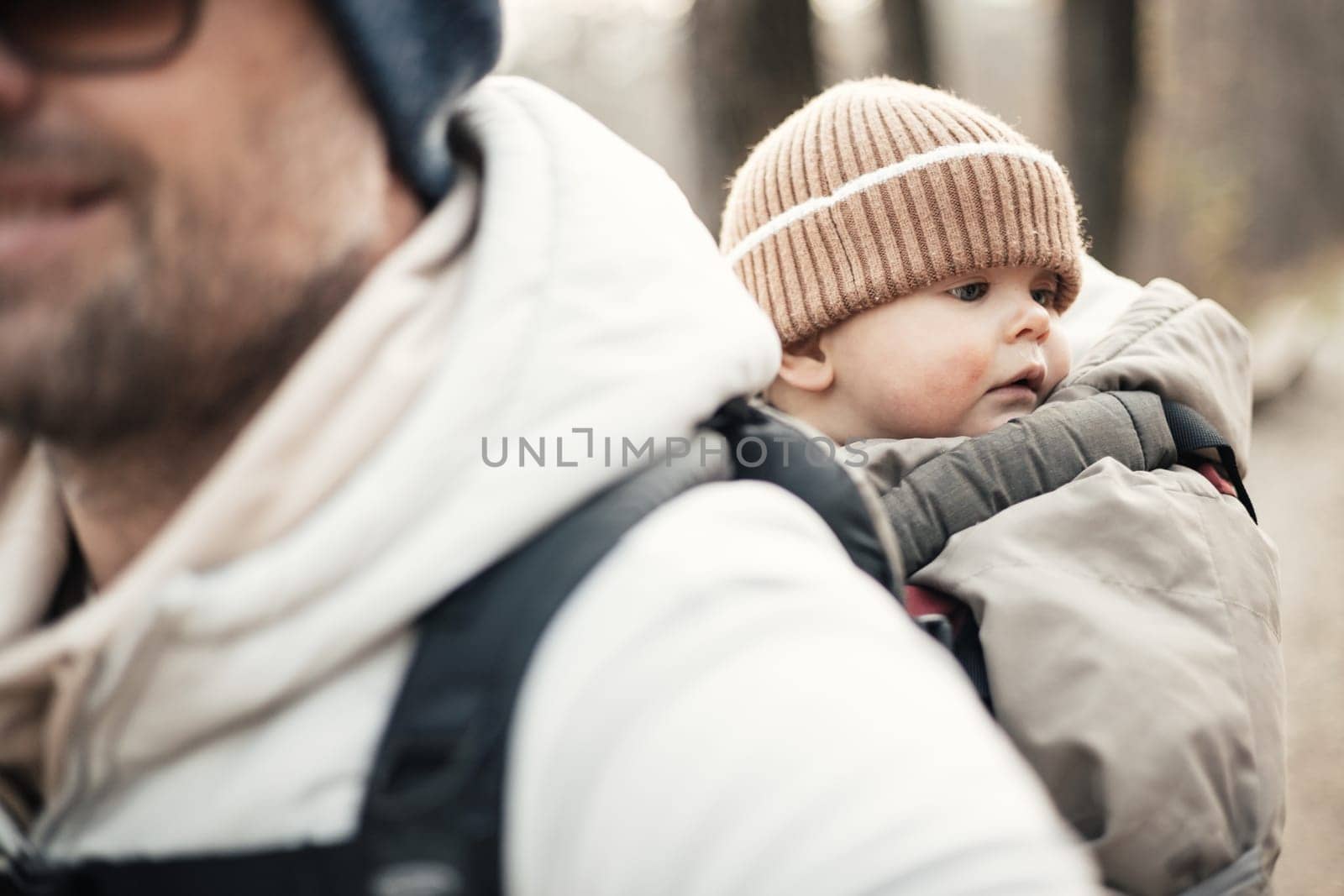 Sporty father carrying his infant son wearing winter jumpsuit and cap in backpack carrier hiking in autumn forest