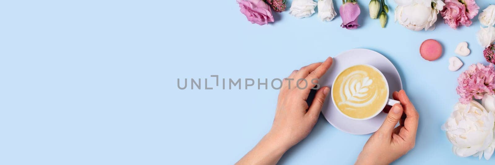 Festive web banner with a blue background. Women's hands hugging a cup of delicious latte macchiato surrounded by roses, peonies, macarons, and pieces of sugar in the form of hearts.