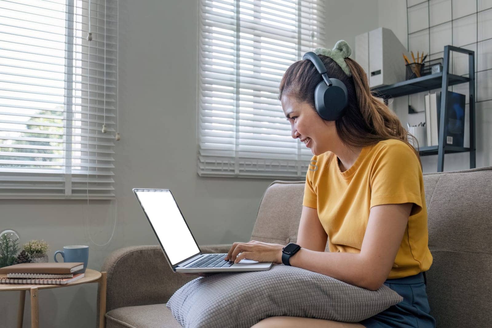 Asian woman sitting on sofa in living room using laptop and listening to music on headphones..