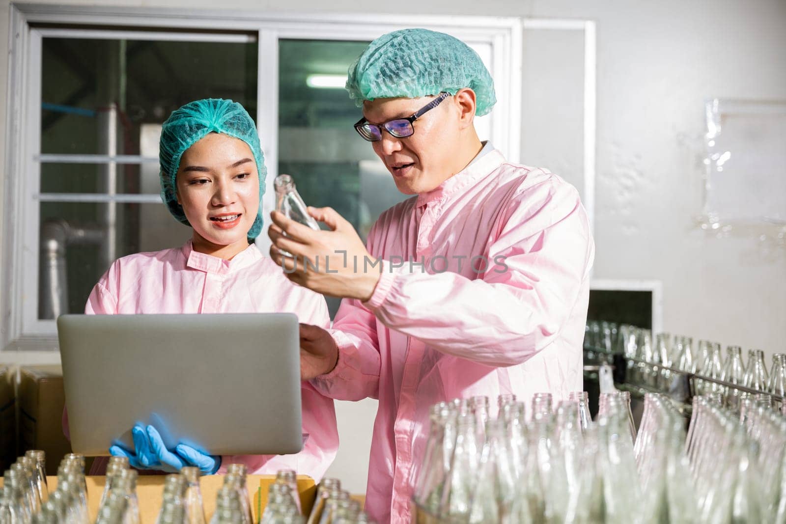 Two engineers conduct detailed inspections on product bottles in a beverage factory's conveyor belt. Quality control and inspection managed by professionals using a laptop.