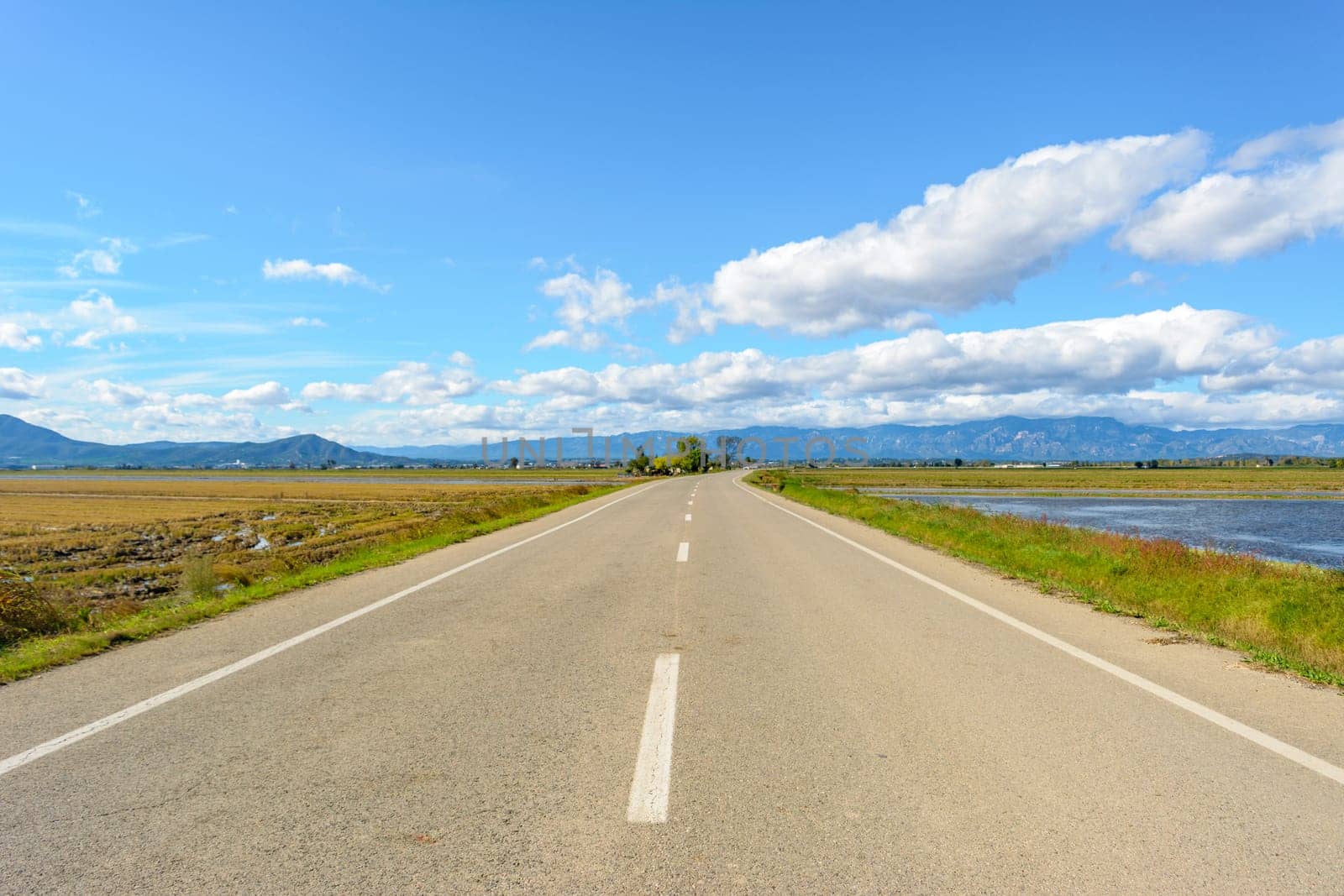 A straight road leading towards mountains under a bright blue sky with clouds, view of lonely road in the ebro delta, tarragona, catalonia, spain,