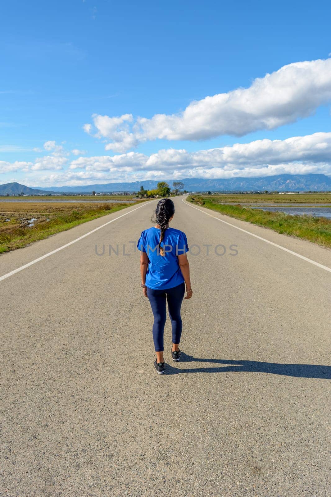 View from behind an individual walking along a deserted road with scenic views on a clear day, back view of latina woman dressed in blue walking down the road in the Ebro Delta natural park, Tarragona, Catalonia, Spain,