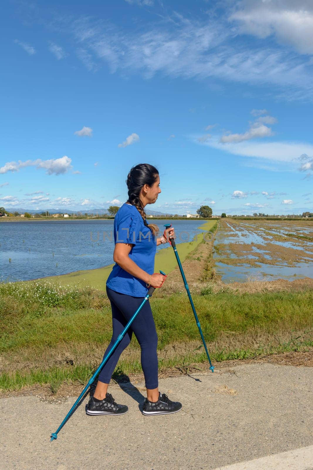 A woman in a blue shirt Nordic walking along a waterway on a sunny day with a clear blue sky, Hispanic latin woman walking with trekking poles in the Ebro Delta natural park, Tarragona, Catalonia, Spain,