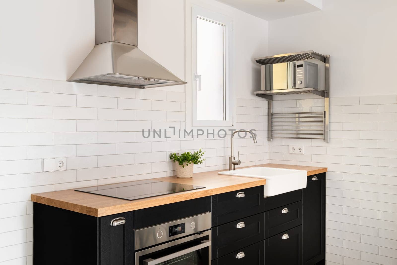 Kitchen island with convection hob and oven with black drawers and wood panel and sink with window and extractor hood against white brick wall. Concept of modern renovation in a new apartment by apavlin