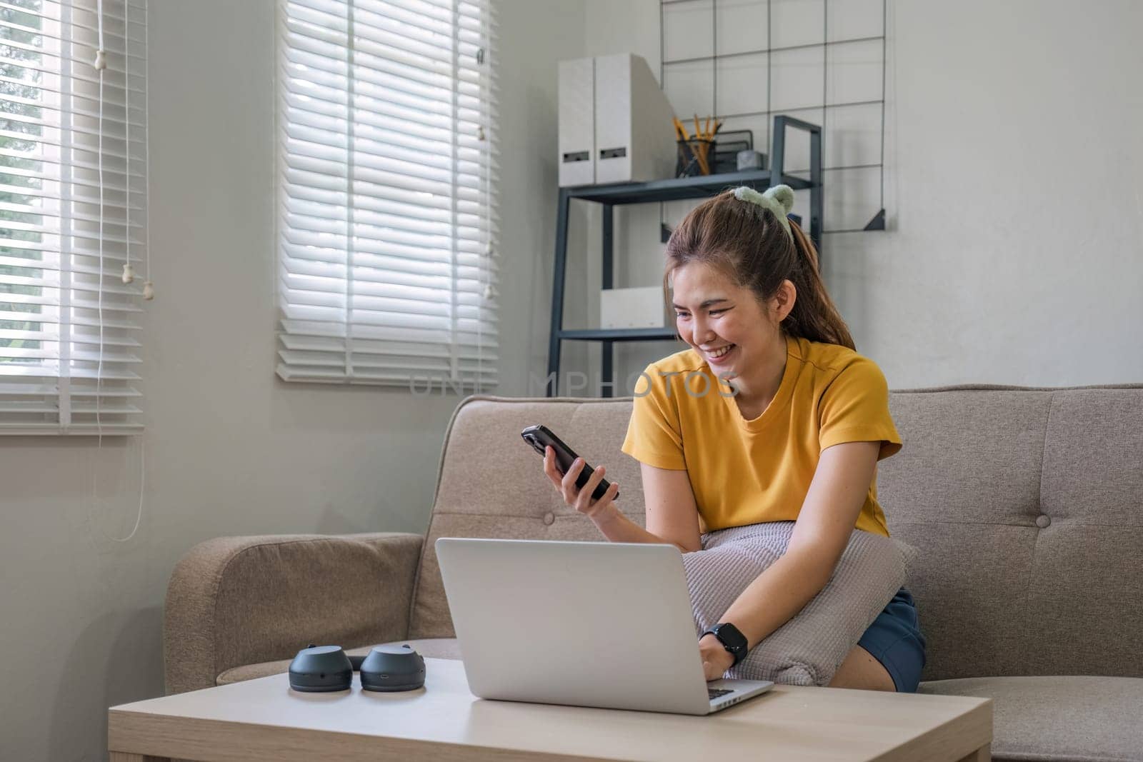 Happy young Asian woman uses mobile phone while sitting on sofa at home with laptop computer..
