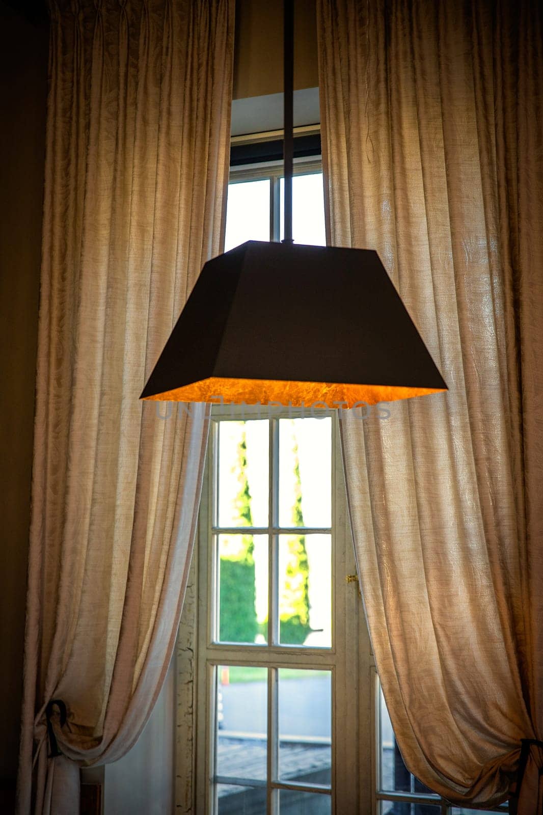 Modern brown lamp hanging from the ceiling shines with warm light in front of a window with curtains.