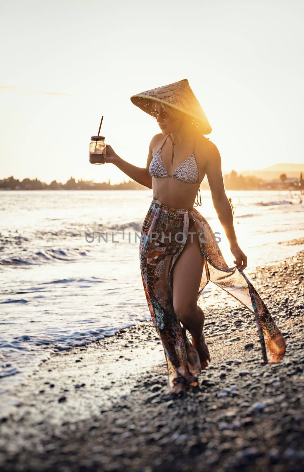 Beautiful girl with a smoothie by the sea in a straw Asian triangular hat on the sunset background by Rotozey