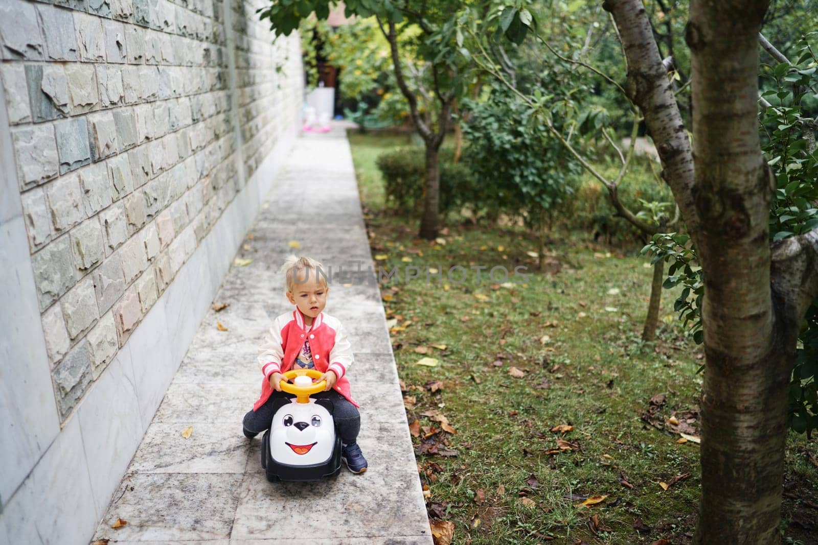 Little girl rides a colorful toy car along the wall in the garden. High quality photo