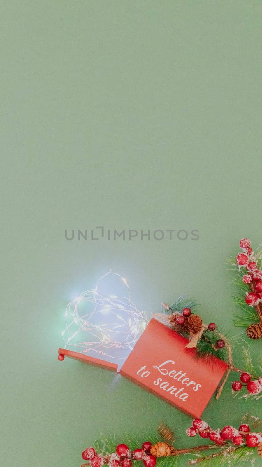 One Santa Claus mailbox, fir branches and a burning garland falling out of it on a pastel green background with copy space, flat lay close-up.