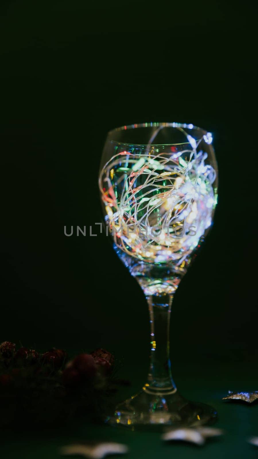 One wine glass with a colored burning garland in it stands at night on a dark green background, close-up side view.