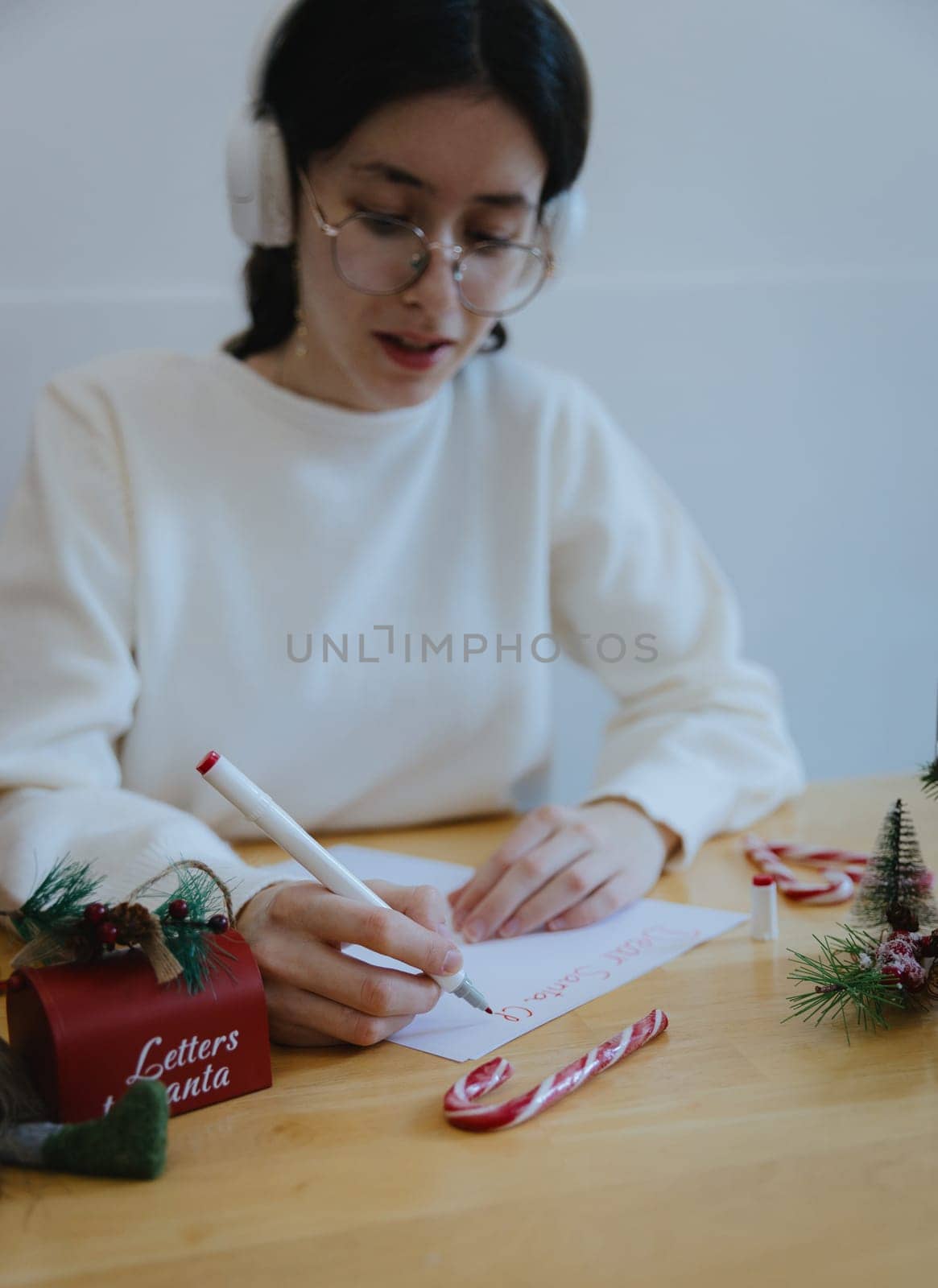 One beautiful recognizable Caucasian teenage girl emotionally writes a letter to dear Santa Claus with a red felt-tip pen, sitting at a wooden table with Christmas decorations, in the dining room on a winter day, side view from above, close-up.