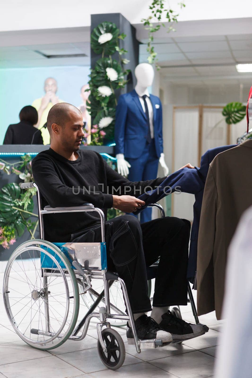 Arab man in wheelchair examining formal jacket in department store fashion showroom. Boutique client with disability browsing through apparel rack while shopping for outfit