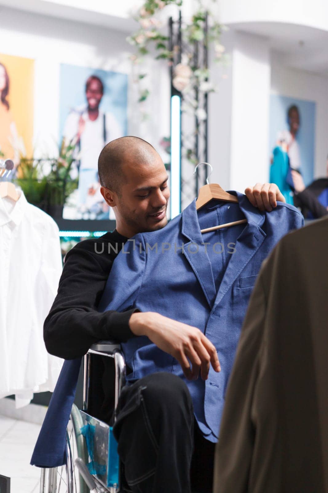Smiling arab buyer in wheelchair holding jacket to body, examining formal apparel fit and size before making purchase in boutique. Clothing store customer with disability choosing outfit