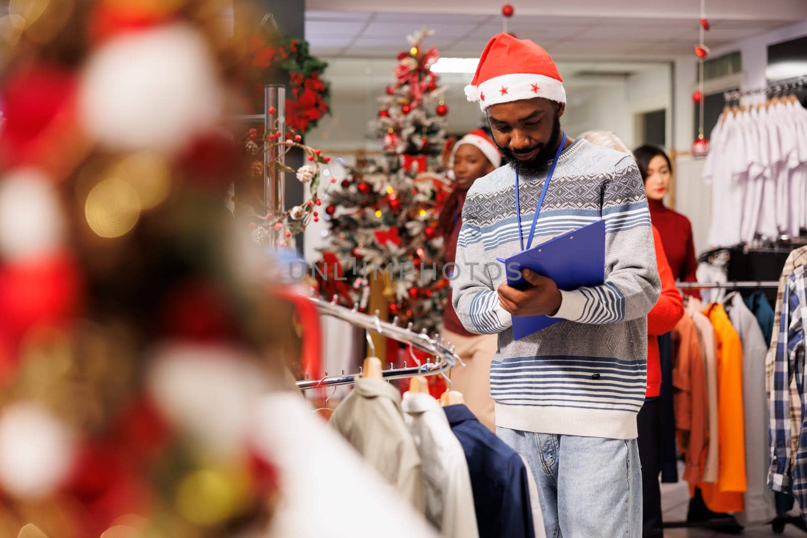 In festive textiles store, sales associate wears santa hat and completes inventory of goods in stock while holding clipboard. Store helper summing every item and arranging clothes.