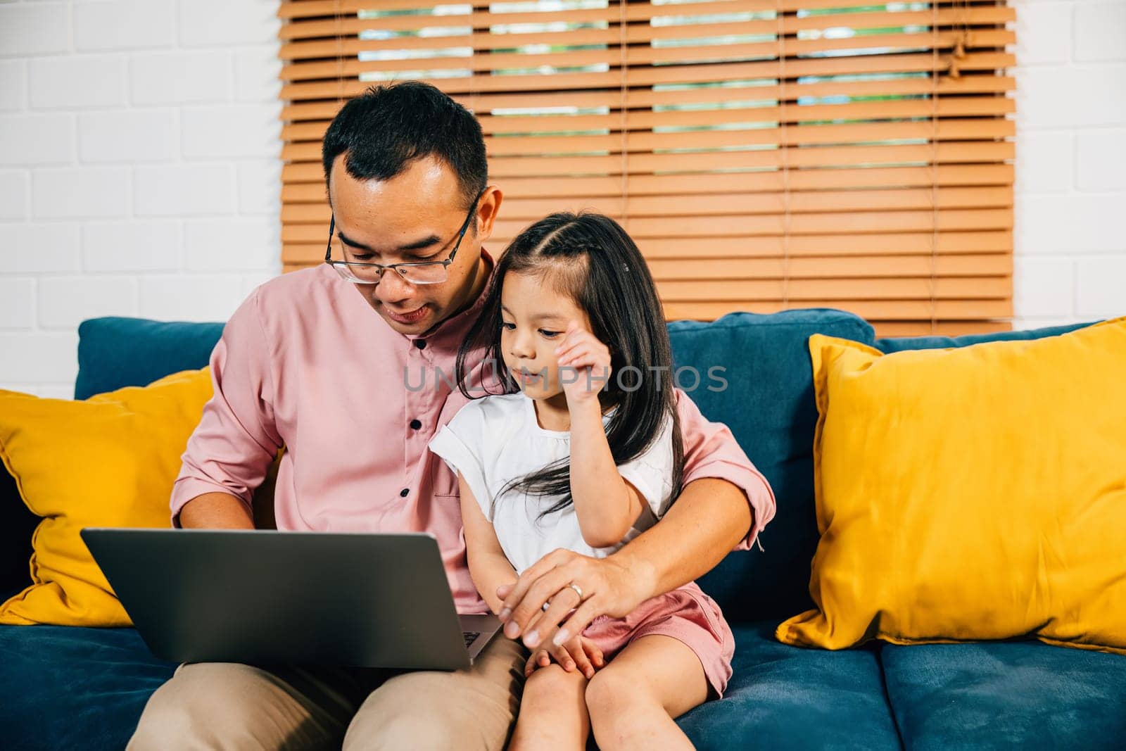 An Asian father finds happiness in his work on a laptop while his daughter enjoys e-learning on a computer in their modern living room. Their togetherness and smiles create a special family moment.