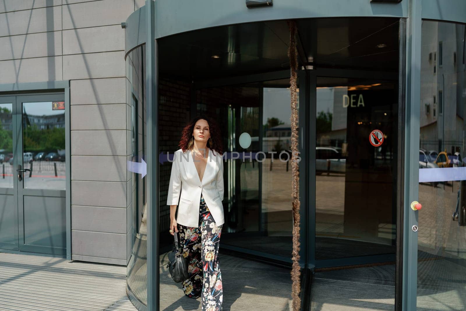 Woman leaves a supermarket. Caucasian model with long dark hair, wearing a white jacket and colored trousers