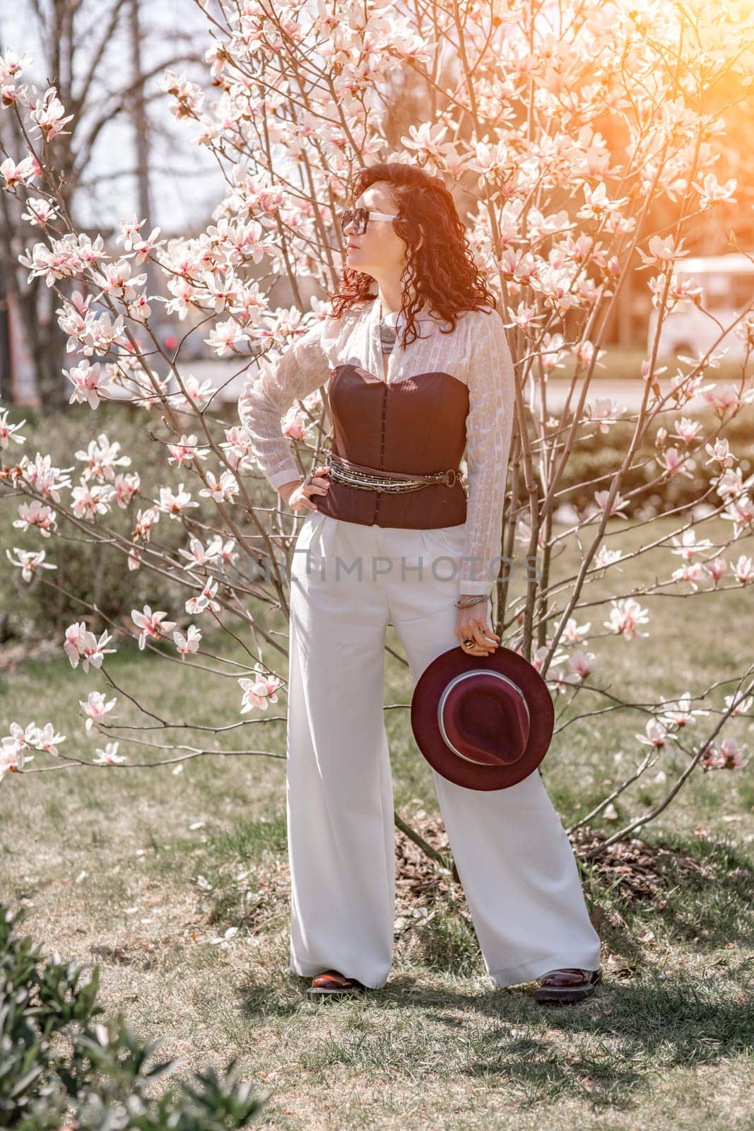 Magnolia park woman. Stylish woman in a hat stands near the magnolia bush in the park. Dressed in white corset pants and posing for the camera. by Matiunina