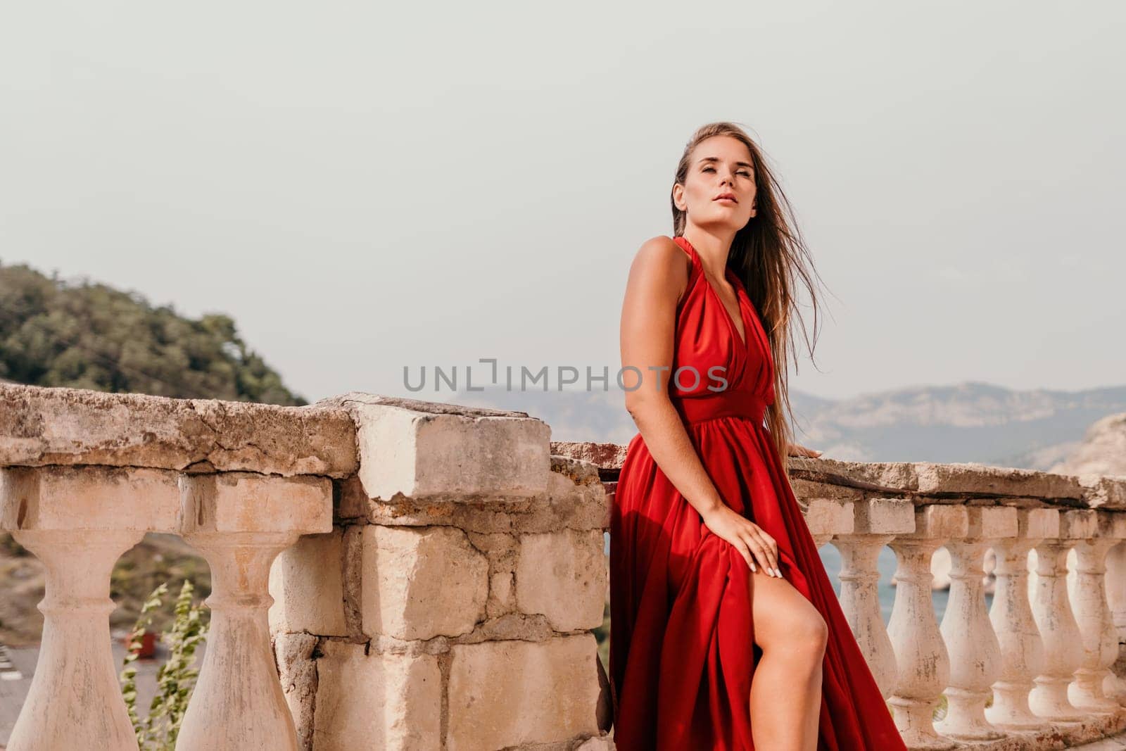 Side view a Young beautiful sensual woman in a red long dress posing on a volcanic rock high above the sea during sunset. Girl on the nature on overcast sky background. Fashion photo