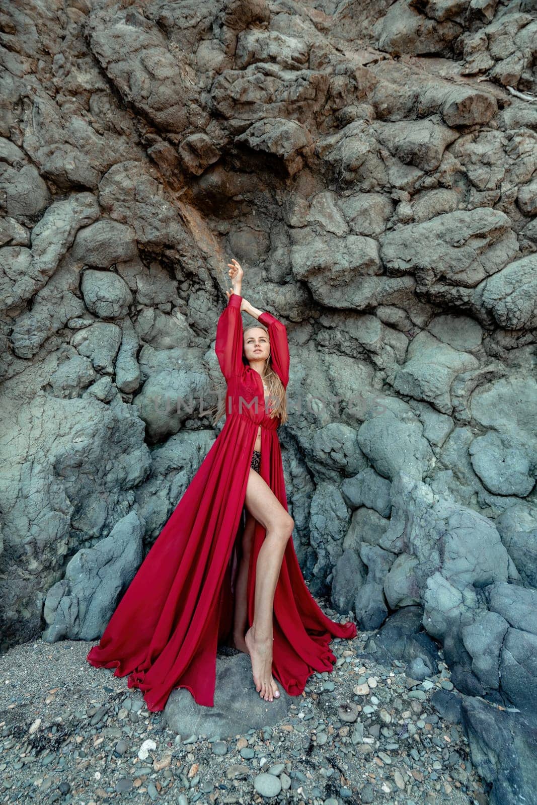 Red dress rocks woman. A blonde with flowing hair in a long flowing red dress stands near a rock of volcanic origin. Travel concept, photo session at sea by Matiunina