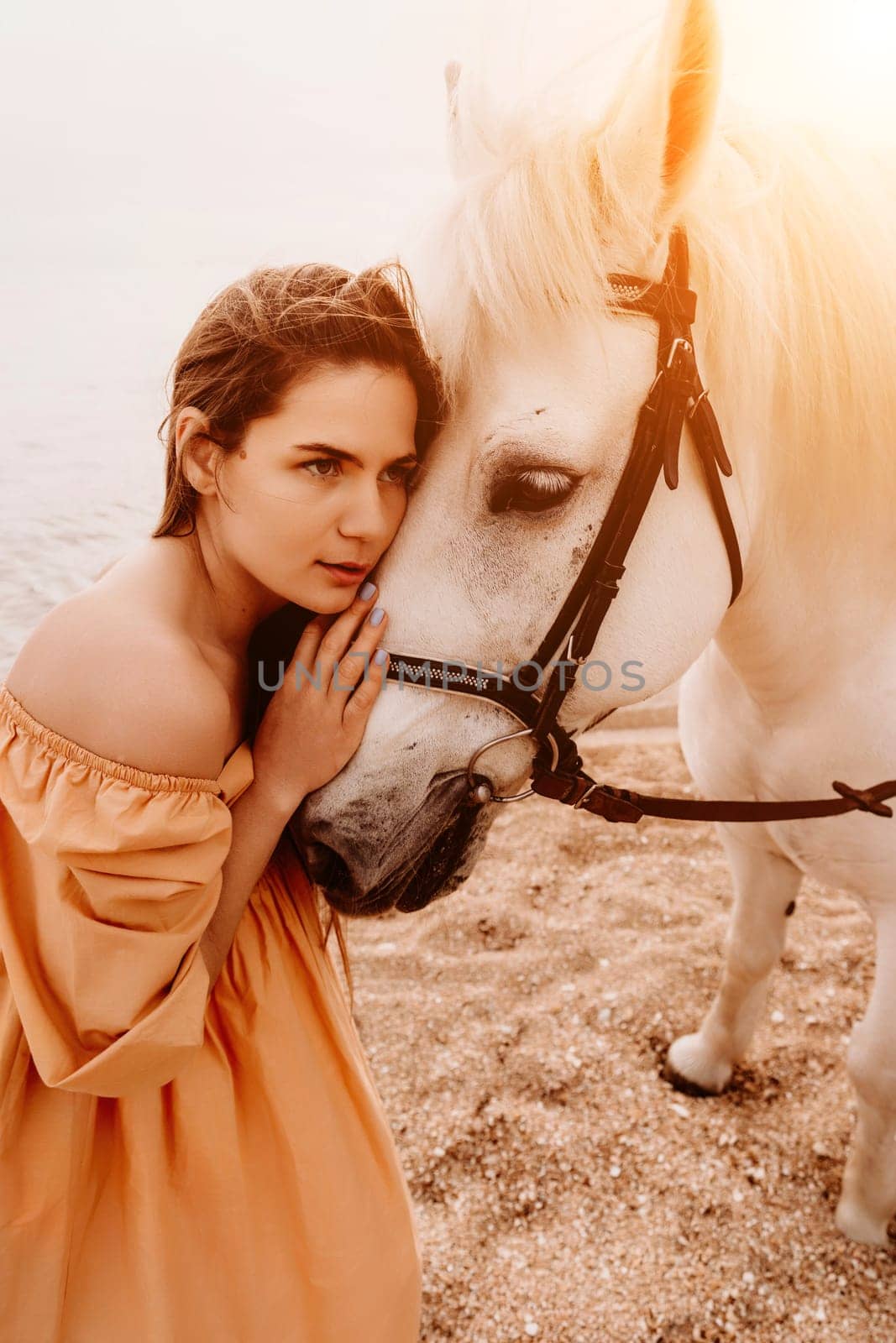 A woman in a dress stands next to a white horse on a beach, with the blue sky and sea in the background