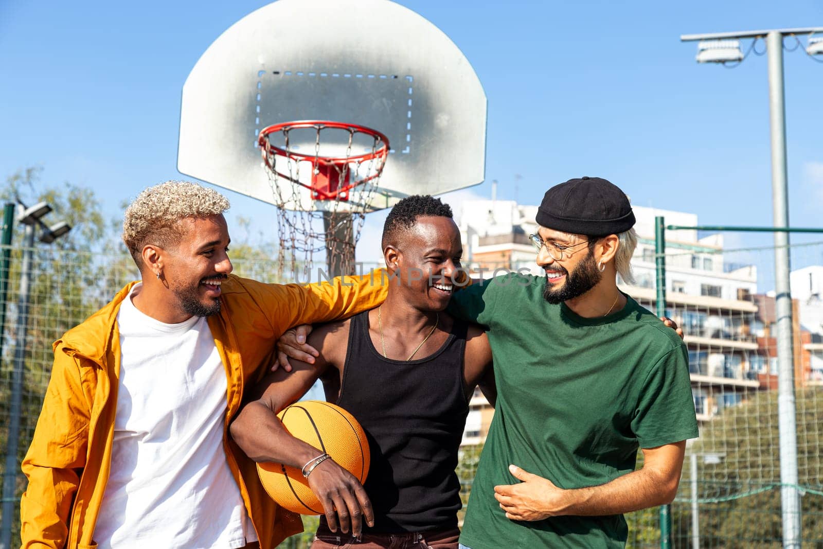 Three happy young multiracial male friends embracing laughing and having fun standing in basketball court outdoors. by Hoverstock