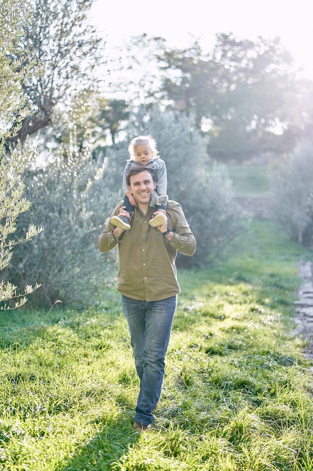 Smiling dad with a little girl on his shoulders walks through the green grass in the park. High quality photo