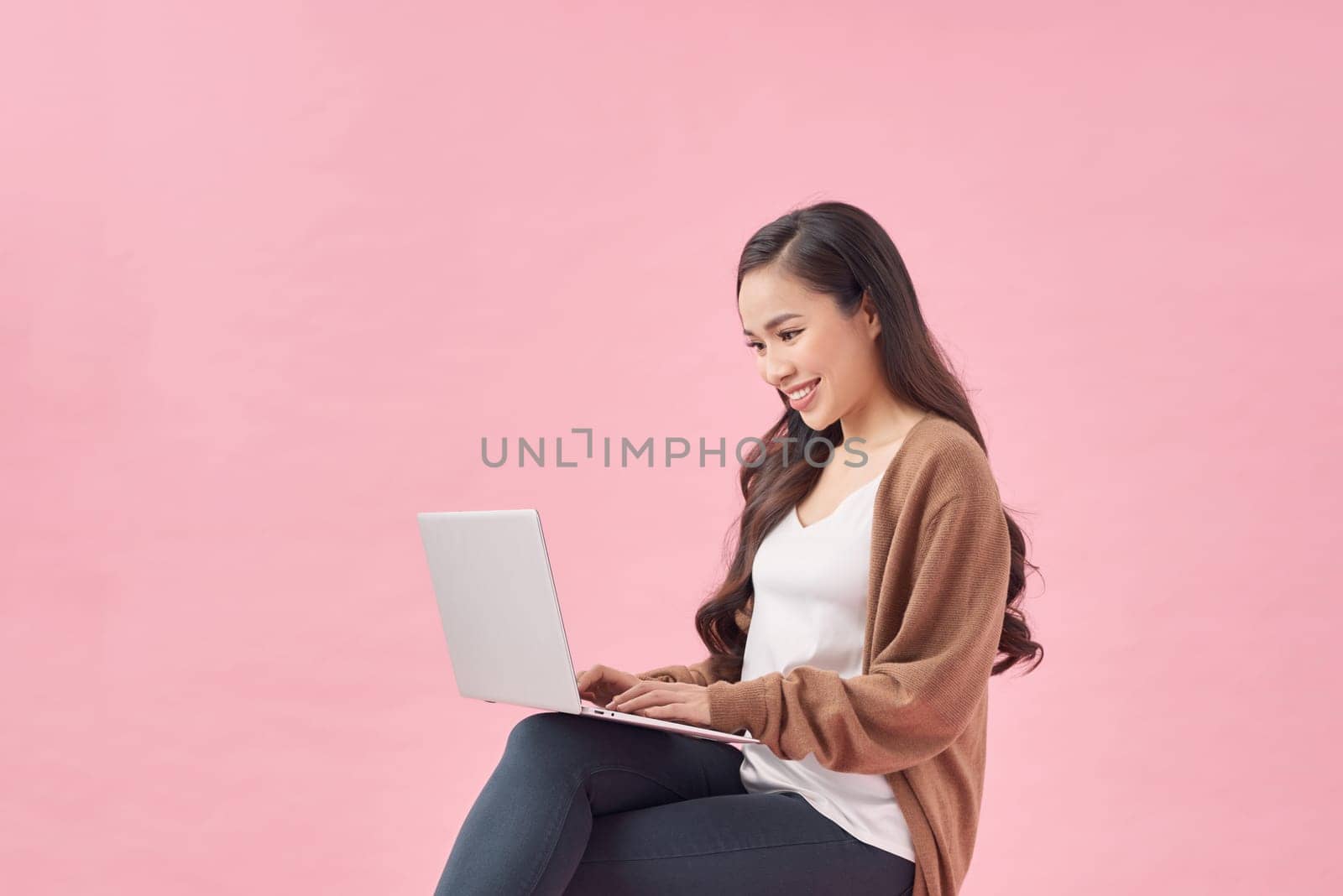 Beautiful young woman sitting on chair, working on laptop against pink wall by makidotvn