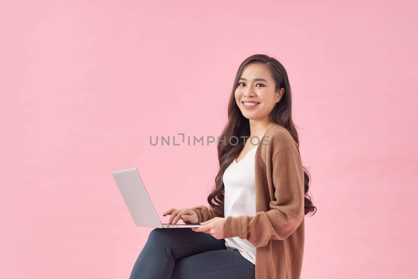Happy young woman working online, sitting on chair and using laptop against pink wall by makidotvn