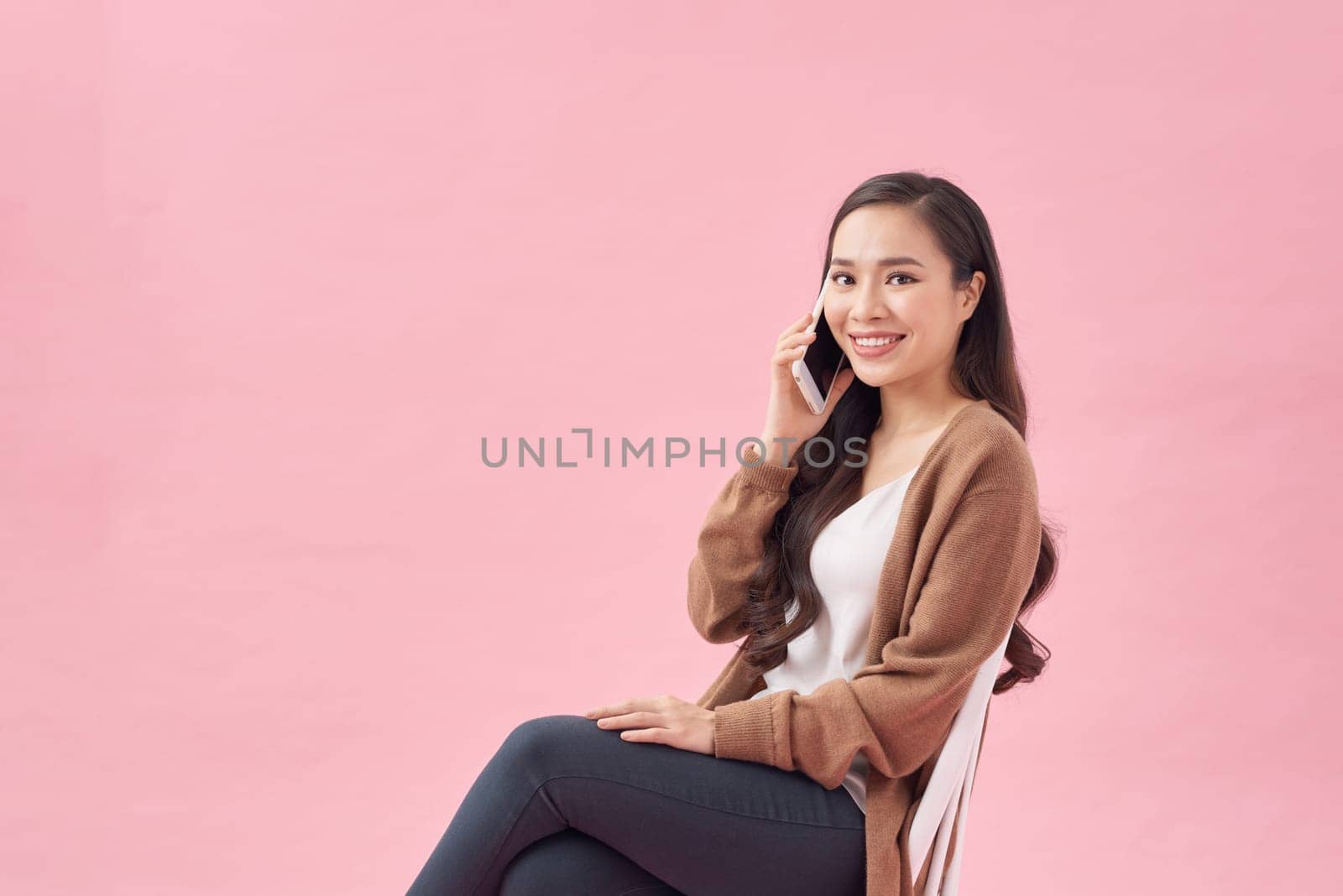 Positive young asian female smiling and talking smartphone while sitting on chair on pink background by makidotvn