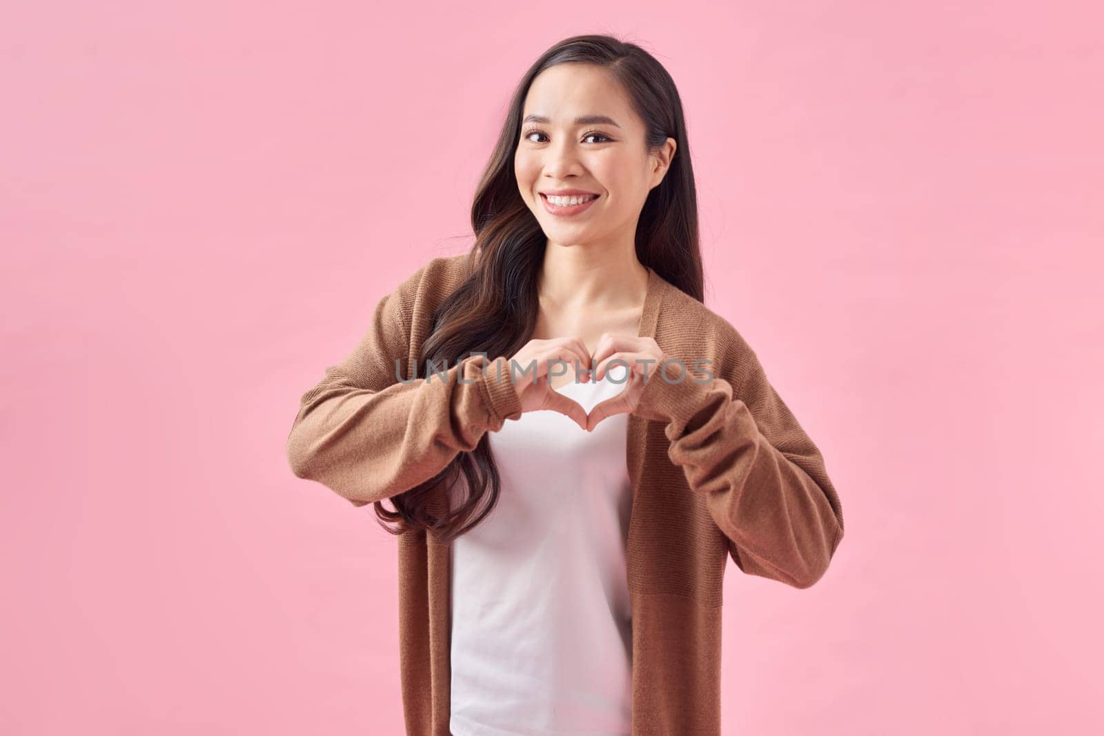 Isolated portrait of beautiful asian woman make heart symbol by hands on pink background