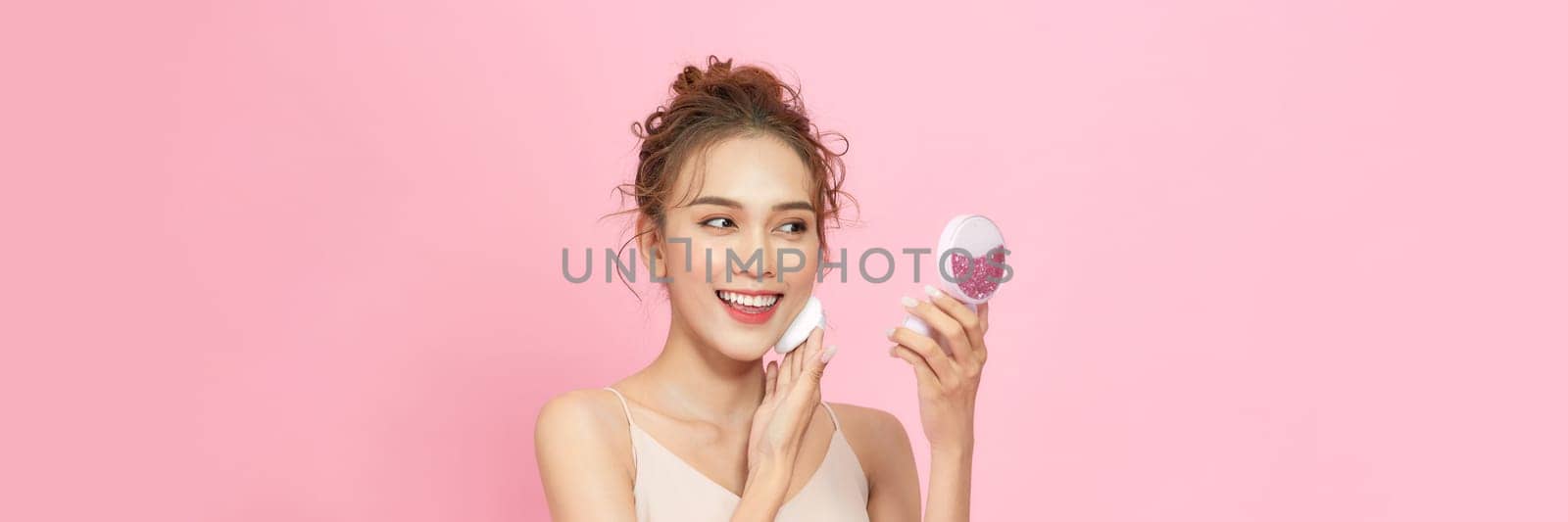 Portrait of attractive cheery girl applying blush tone foundation isolated over pink pastel background by makidotvn