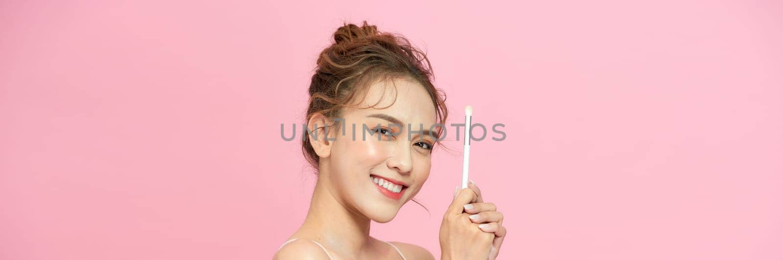 Woman applying face powder on nose making makeup using cosmetic brush posing over pink by makidotvn